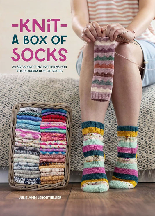 Cover of “Knit A Box Of Socks” by Julie Ann Lebouthillier, featuring a person wearing colorful knitted socks and holding another sock in progress. A basket filled with vibrant sock pairs sits nearby. The subtitle reads “24 Sock Knitting Patterns For Your Dream Box Of Socks.”