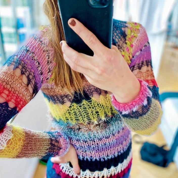 Mirror selfie of a person wearing a vibrant, multicolored knitted sweater featuring eclectic patterns in pink, purple, blue, yellow, and green. The person’s face is partially hidden by the phone, highlighting the bold and expressive knitwear design.