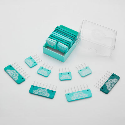 A set of blue and teal rectangular knitting blockers  with metal prongs, some inside a clear plastic box and others arranged outside on a white surface.