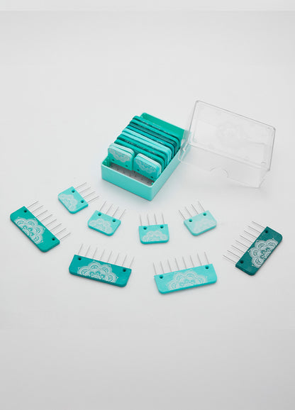 A set of blue and teal rectangular knitting blockers  with metal prongs, some inside a clear plastic box and others arranged outside on a white surface.