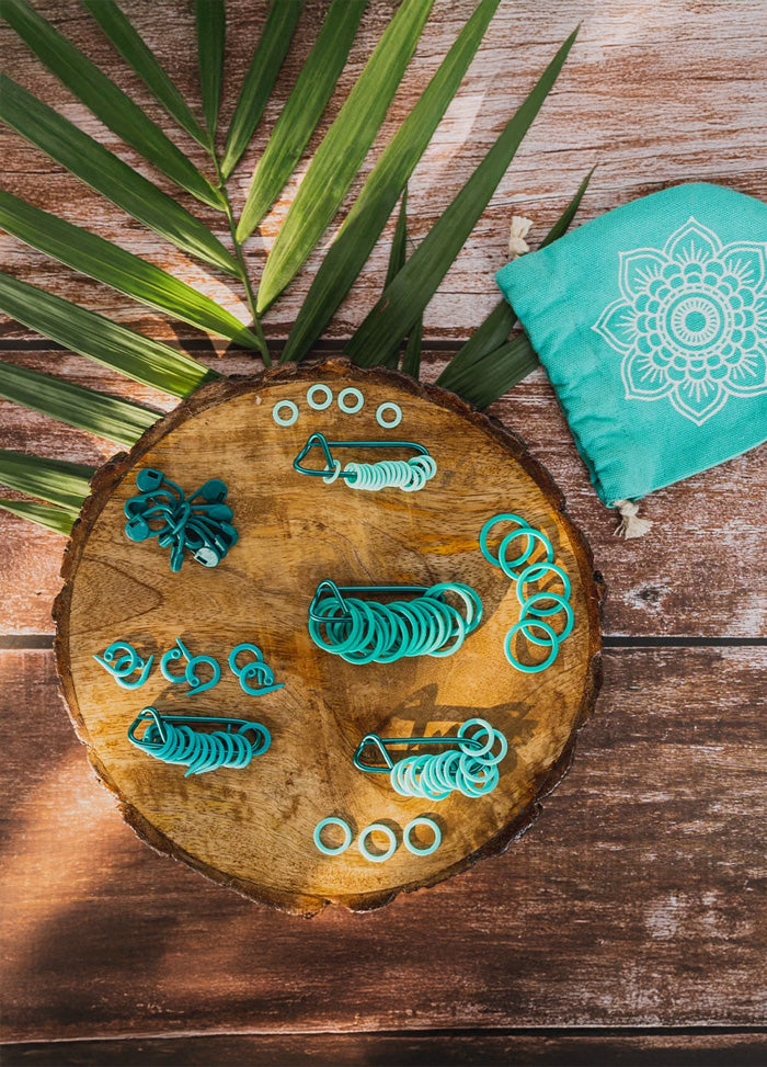  On a wooden table, a green bag holds teal stitch markers, with blue rubber bands arranged nearby for easy access.