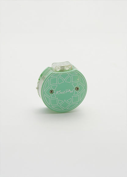 Row counter with a flower design in green and white, used for tracking rows on a teal knitting tool in a cotton pouch