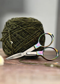 Brightly colored folding scissors with a metallic sheen placed next to a ball of yarn, emphasizing their stylish and safe design.