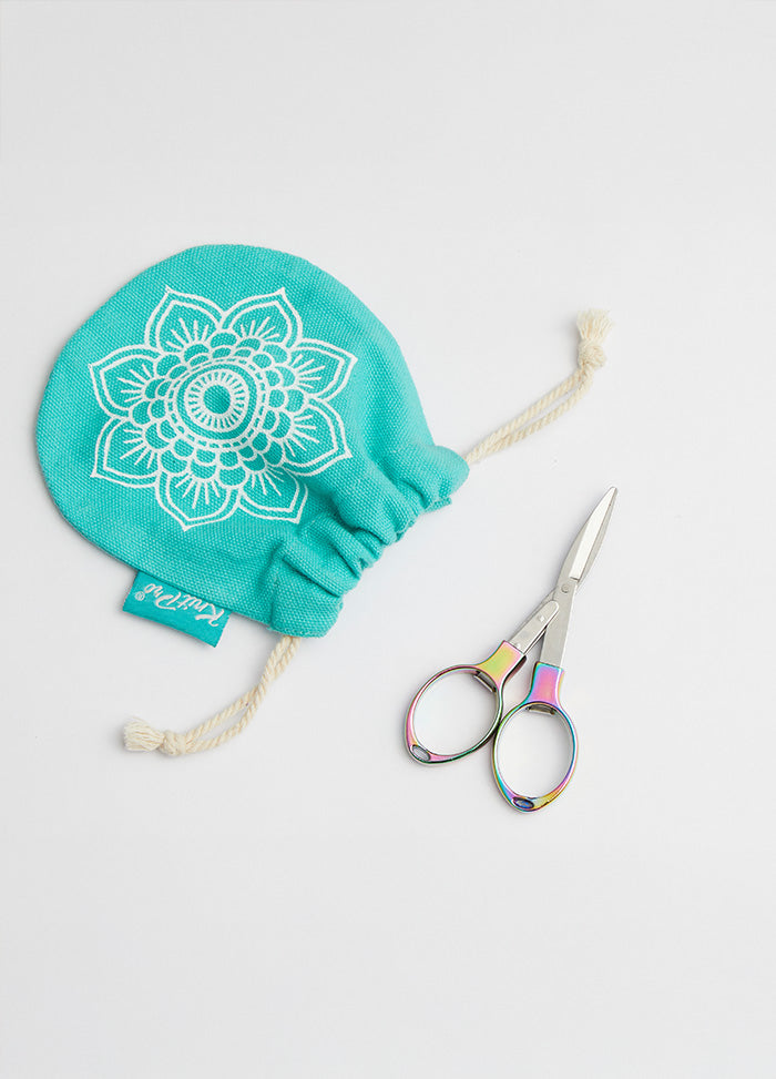A pair of colorful, foldable scissors with a shiny finish next to a bag adorned with a floral design.