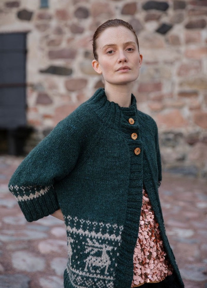 Hiiden Hirven Hiihto: dark green cardigan with deer motif near the hem and wooden buttons, set against a stone wall and cobblestone ground.