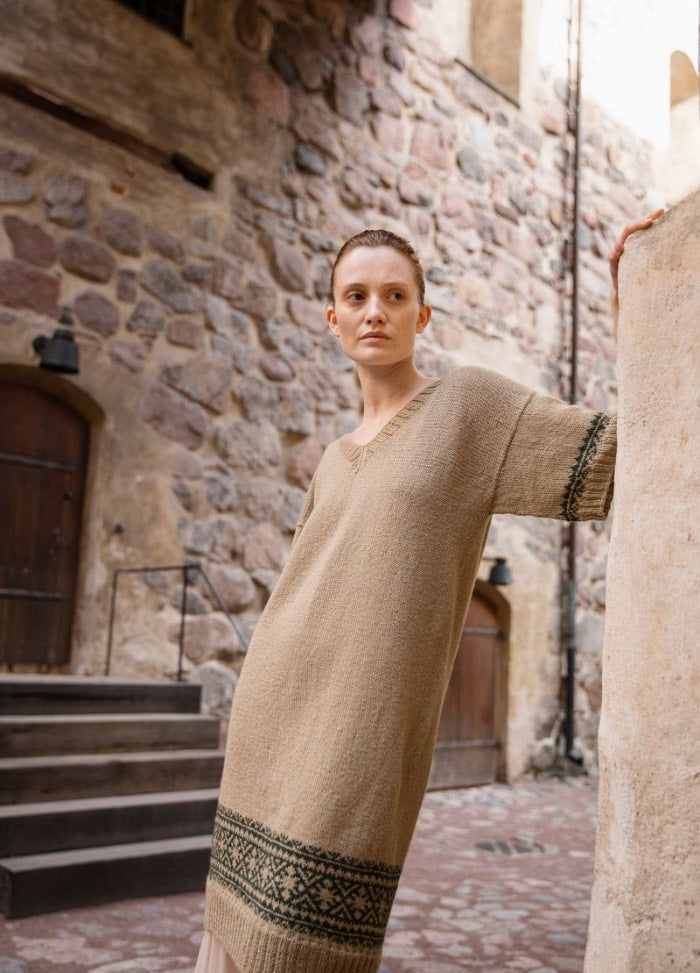 Kaitse Karja Kaunihisti: beige knitted dress with green and black sleeve and hem motifs, photographed outdoors near stone walls and wooden doors.