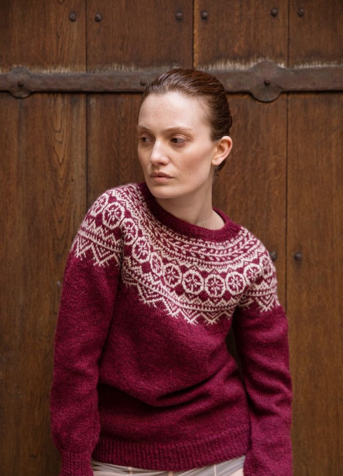 Kotimatka: maroon sweater with white geometric and snowflake-style yoke patterns, set against a wooden paneled backdrop with metal hinges.