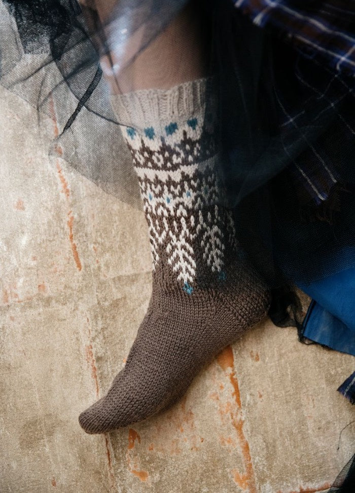 Kullervo: multicolored sock in brown, white, and blue with intricate patterning, photographed on a textured floor surface.