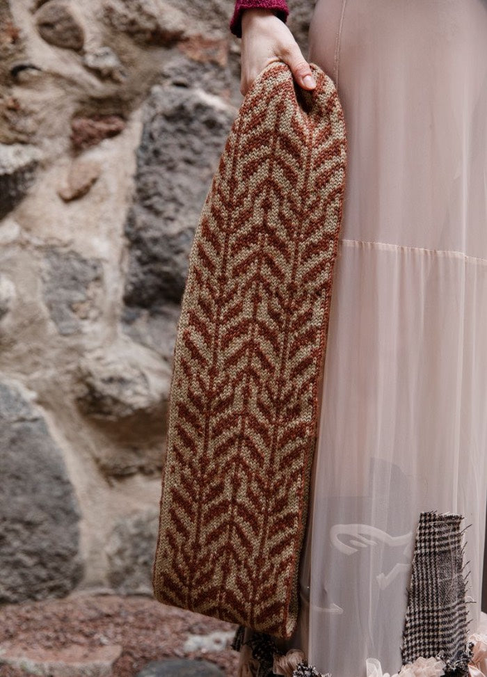 Kuunteleva Metsa: brown and beige cowl with symmetrical interlocking motifs, held against a textured stone wall.