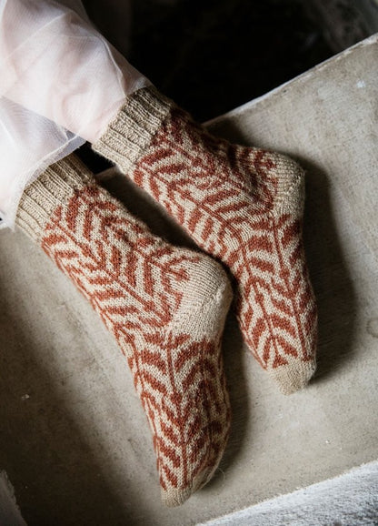Kuunteleva Metsa: beige socks with red-brown leaf motifs, worn with sheer fabric and photographed on a stone surface.