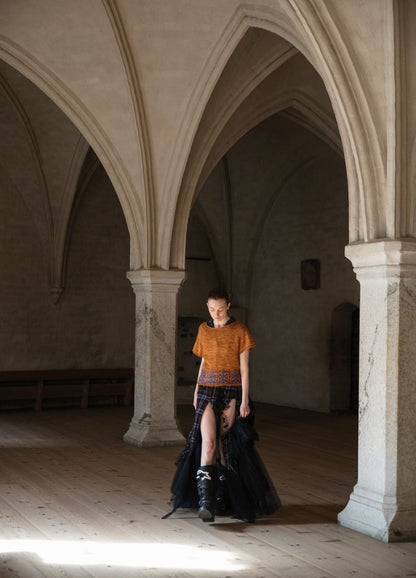 Lahde Kukka Kulkemahan: orange top paired with a black patterned skirt, photographed in a grand arched stone interior.