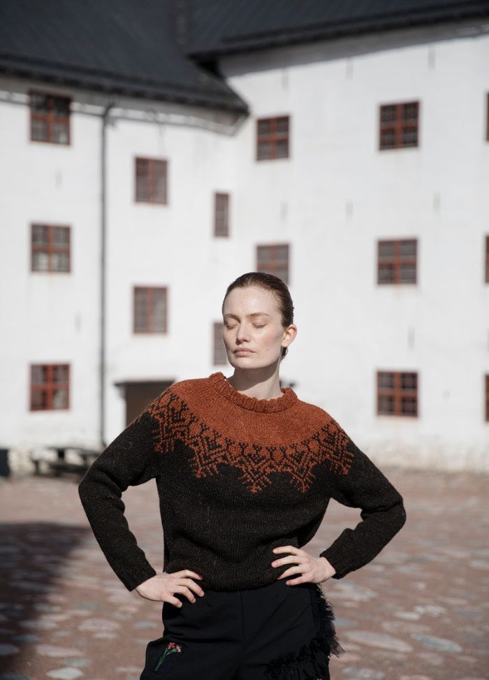 Lemminkainen: dark sweater with bold orange shoulder and neckline motifs, photographed near a white building with cobblestone pavement.