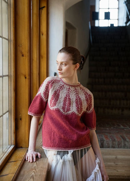 Meren Luo'olle Lokiksi: red and white sweater with intricate motifs, photographed beside a wooden window frame with light streaming in.