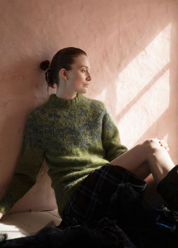 Mull' On Korvessa Kotini: green sweater with blue motifs, paired with a dark plaid skirt, photographed in sunlight against a light pink wall.