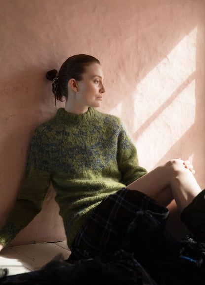 Mull' On Korvessa Kotini: green sweater with blue motifs, paired with a dark plaid skirt, photographed in sunlight against a light pink wall.