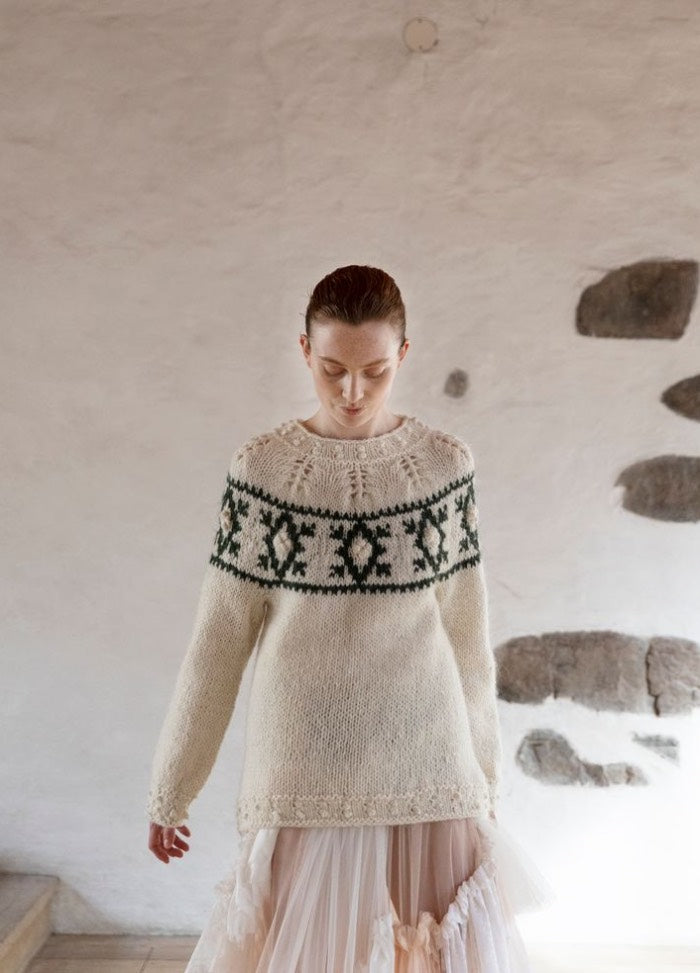Pohjolan haat: cream sweater with green and black geometric yoke pattern, photographed indoors against a stone-accented wall.