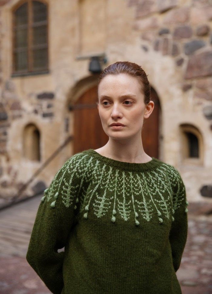 Puiden Synty: green sweater with light green yoke pattern, photographed in front of a historic stone building with arched features.