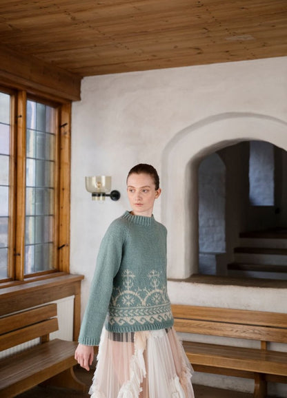 Sariola: light green sweater with white motifs on the waist and cuffs, photographed indoors with wooden benches and arched windows.