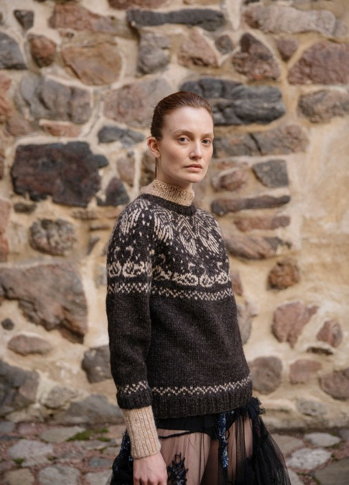 Tuonelan Virta: dark sweater with light intricate motifs on the yoke, cuffs, and hem, set against a multicolored stone wall.