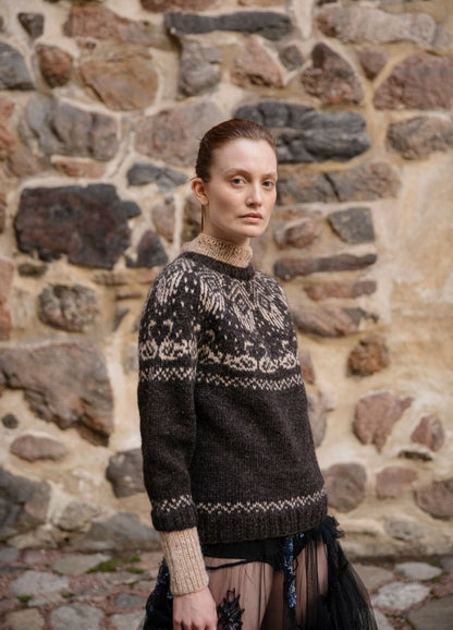 Tuonelan Virta: dark sweater with light intricate motifs on the yoke, cuffs, and hem, set against a multicolored stone wall.