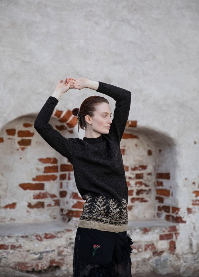 Tuonen Tytti: dark sweater with detailed lower motifs, photographed against a rustic brick wall with arched niches.