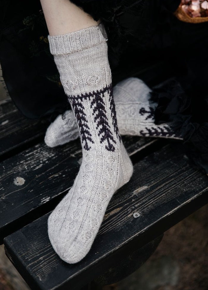 Vellamo: light gray socks with dark vertical motifs and textured knit, worn while seated on a rustic wooden bench.