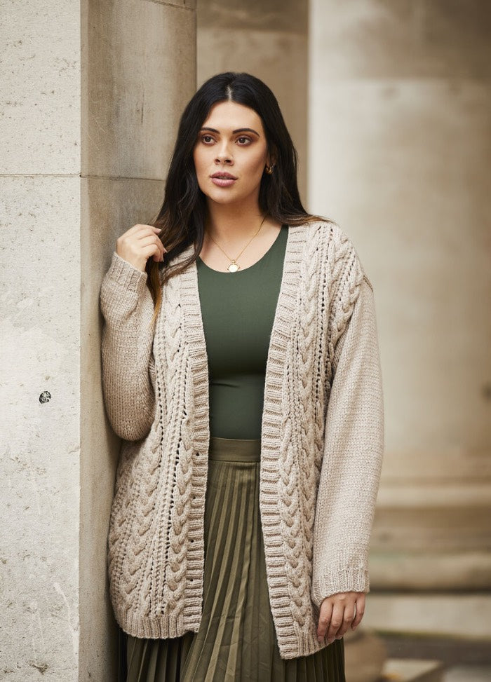 A woman with long dark hair leans against a stone wall, wearing a beige knitted cardigan, olive green dress, and a gold necklace. She appears calm and thoughtful.