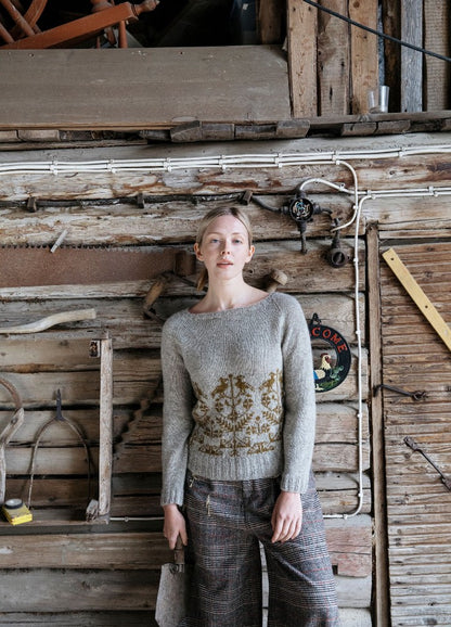 Kultainen K?§ki Sweater: grey knit with a golden motif around the waist, styled with plaid pants against a rustic wooden backdrop.