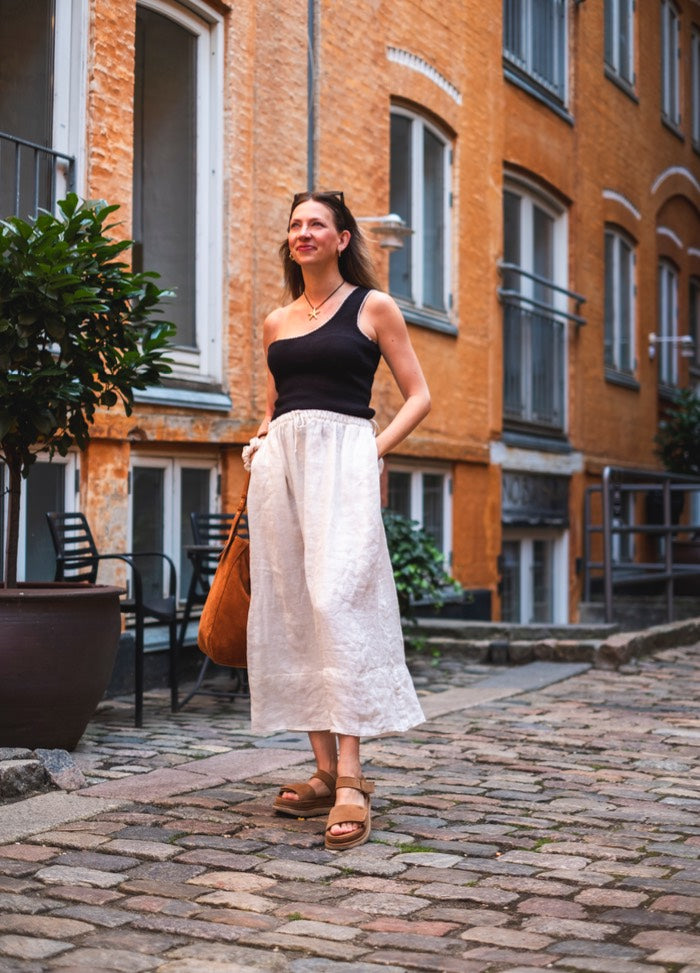 Fortuna Top: Black sleeveless knit styled with white pants and sandals, set against a brick building on a cobblestone street.