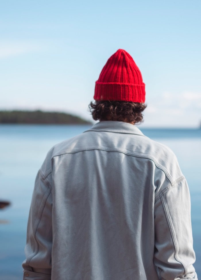 Keep It Together Beanie: Red knit hat worn by the water, styled with a light jacket in a serene outdoor setting.