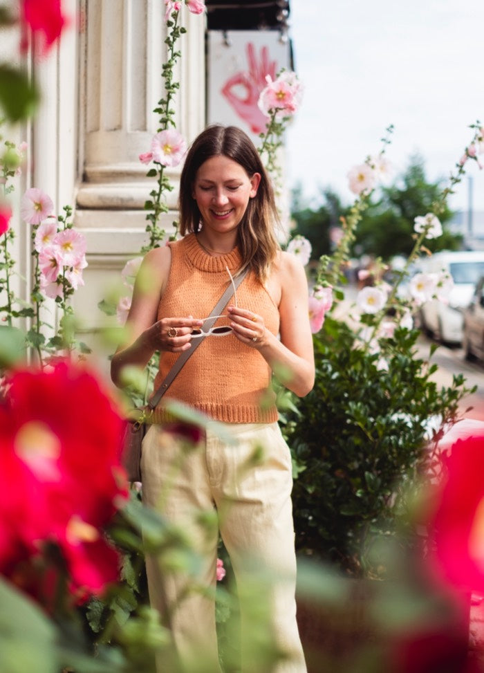 Sundazed Top: Sleeveless orange knit styled with light pants and a crossbody bag, set among blooming flowers.