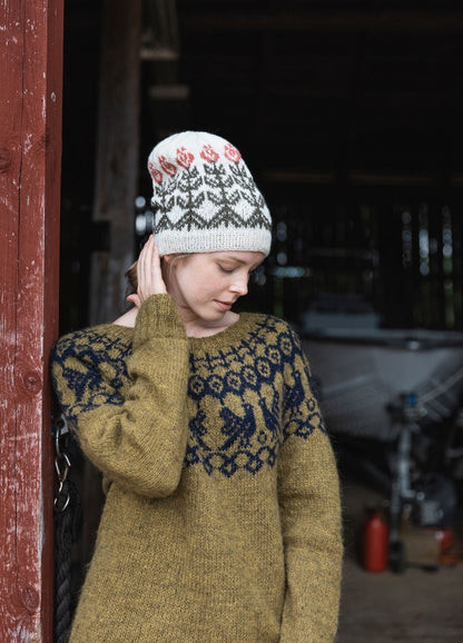 Kyllikki Hat: white knit hat with black and red motifs, worn with an olive green sweater beside a rustic wooden structure.