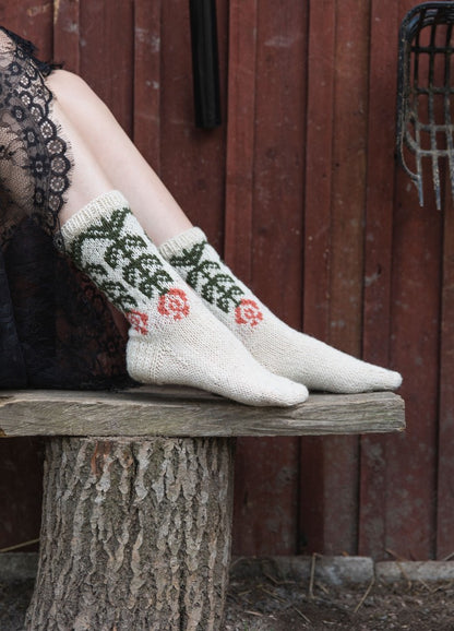 Kyllikki Socks: white knit socks with green leafy and red floral motifs, worn with black lace against a rustic wooden backdrop.