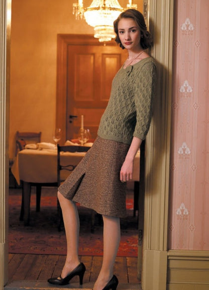 A woman leans against a doorframe, wearing a green knit sweater and brown skirt. The room has a warm, elegant ambiance with a chandelier and set dining table.