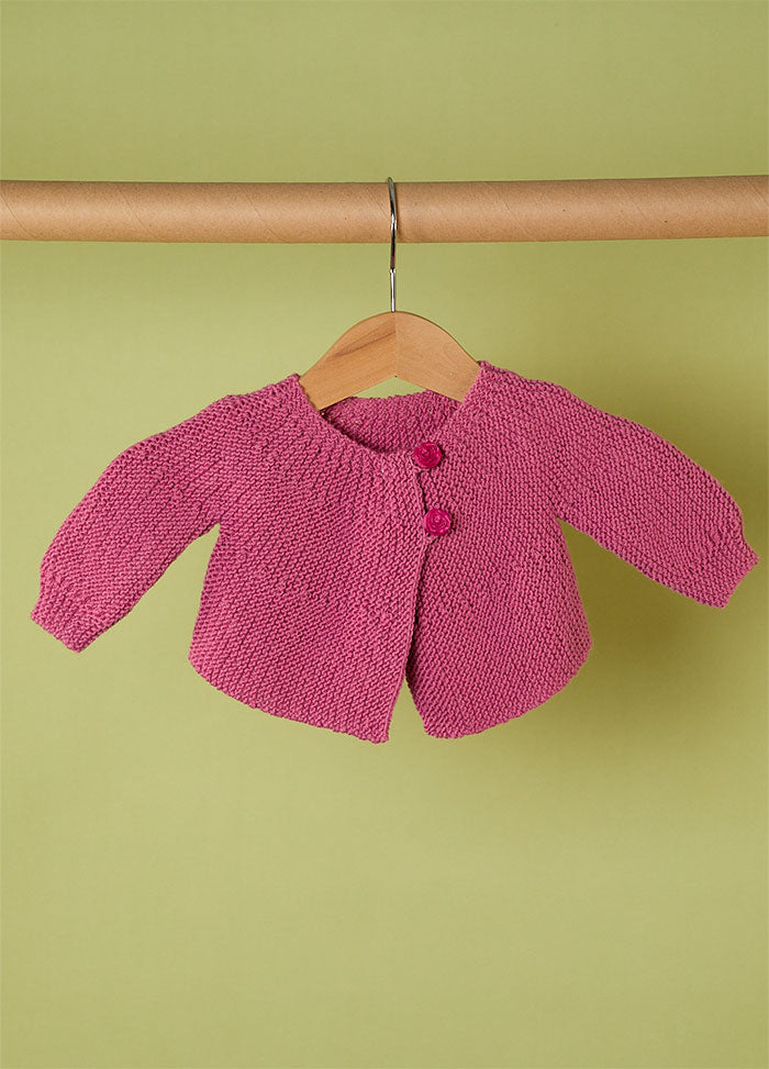 Pink hand-knitted baby cardigan with long sleeves and two front buttons, displayed on a wooden hanger against a light green background – soft and elegant handmade babywear.