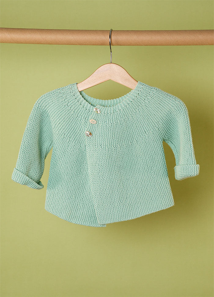 Light green hand-knitted baby cardigan with rolled cuffs and three small buttons near the collar, displayed on a wooden hanger against a matching green background – soft and stylish handmade babywear.
