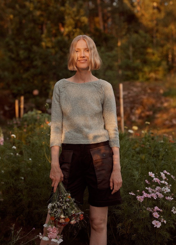 Lucienne Sweater: light-colored knit sweater worn with dark shorts, photographed in a garden setting with flowers and greenery.