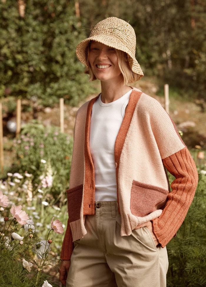 Imatra Cardigan: orange and beige knit cardigan with front pockets, worn in a garden setting over a white shirt and beige pants.