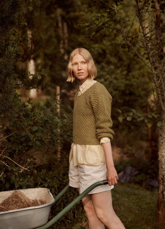 Brisa al Atardecer Pullover: green textured knit sweater worn over a light shirt and white shorts, shown in a garden setting with a wheelbarrow.