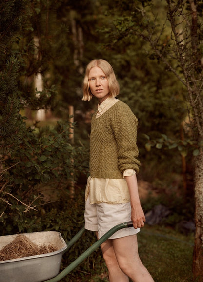 Brisa al Atardecer Pullover: green textured knit sweater worn over a light shirt and white shorts, shown in a garden setting with a wheelbarrow.