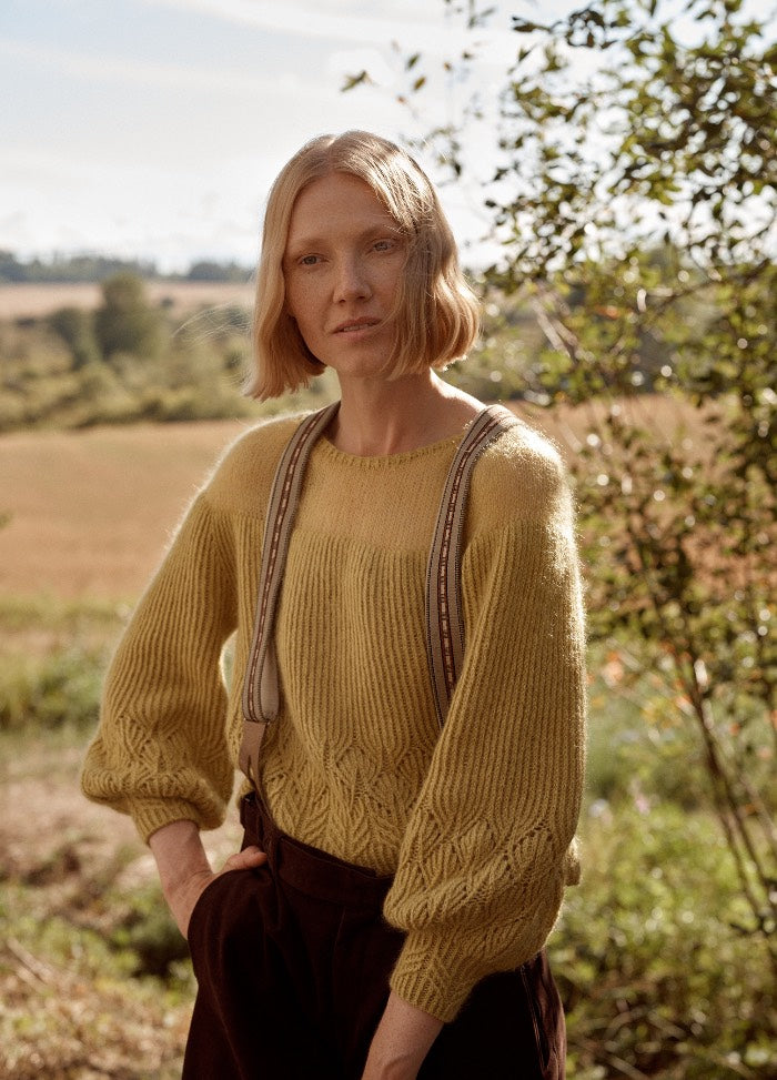 Quince Sweater: yellow knit sweater with detailed sleeve and hem patterns, worn with suspenders in a natural outdoor setting.