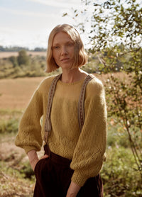 Quince Sweater: yellow knit sweater with detailed sleeve and hem patterns, worn with suspenders in a natural outdoor setting.