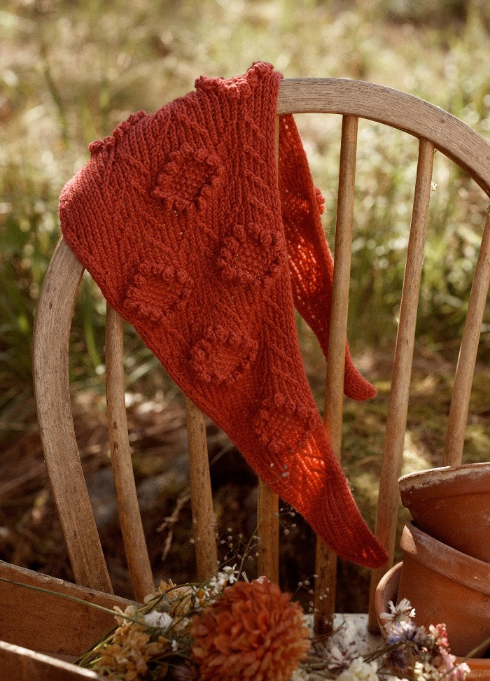 Tsinnia Shawl: red textured knit shawl with raised patterns, draped over a wooden chair in a garden setting.