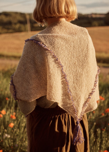 Ziggy Shawl: beige knit shawl with purple fringe and tassel, worn outdoors with fields and flowers in the background.