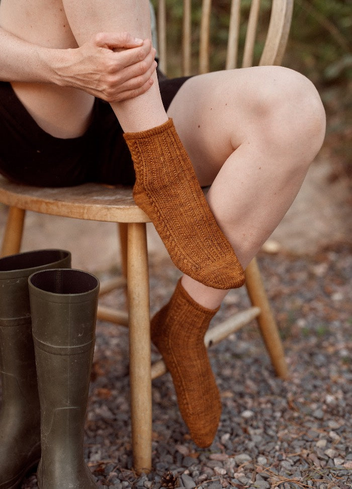 Speculaas Socks: brown knitted socks worn outdoors, with green boots nearby on gravel beside a wooden chair.