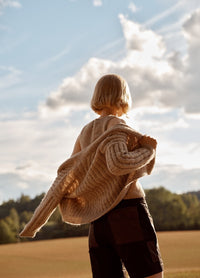Casadinho Cardigan: beige knit cardigan worn in a field setting, shown from the back with a clear sky and trees in the background.