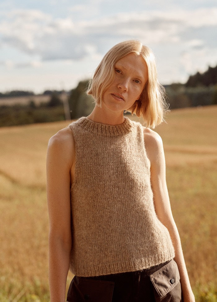 Casadinho Slipover: sleeveless knitted top worn in a field with trees and a partly cloudy sky in the background.