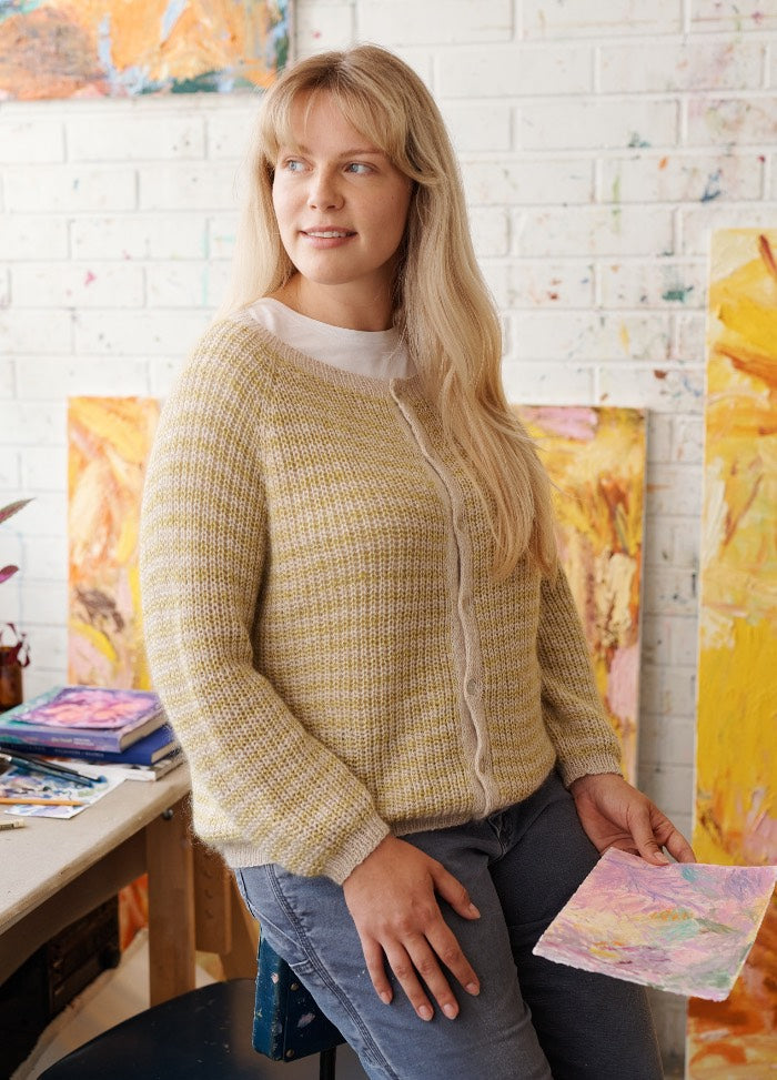 Reversi Cardigan: light yellow and white knit cardigan worn over a white shirt, seated in an art studio with abstract paintings.