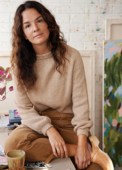 Hiekassa Sweater: beige knit sweater worn with brown pants, seated in an art-filled setting.