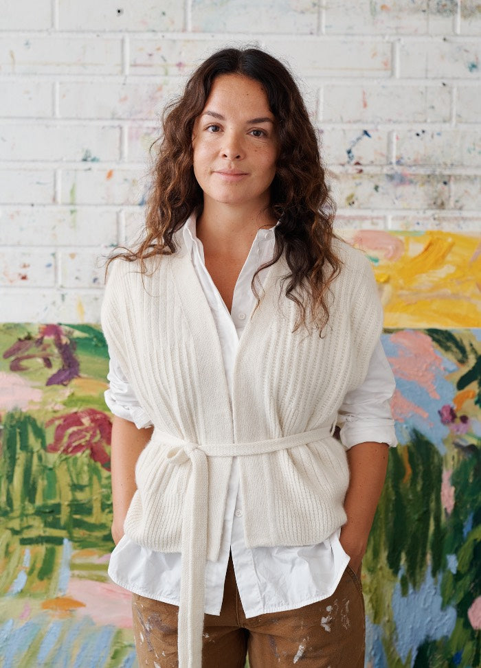 Valo Vest ‚Äì cream knit vest tied at the waist over a white shirt, with colorful abstract art in the background.
